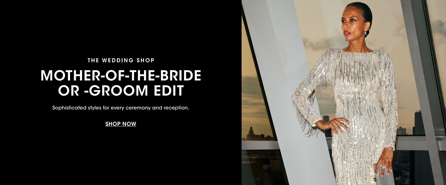 1 photo, of an older female model standing in front of a floor to ceiling window at dusk, one arm on her hip, wearing a sparkling silver long sleeve gown.