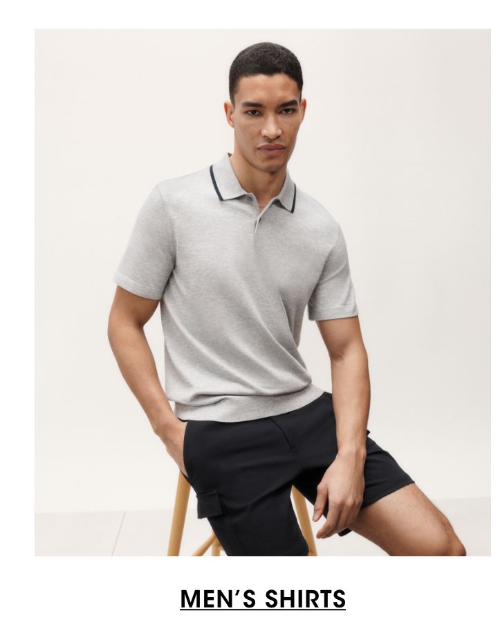 1 photo, of a male model sitting on a stool in front of a plain white background. Model looks directly at the camera, one hand in his pocket, wearing a gray polo shirt and black shorts.