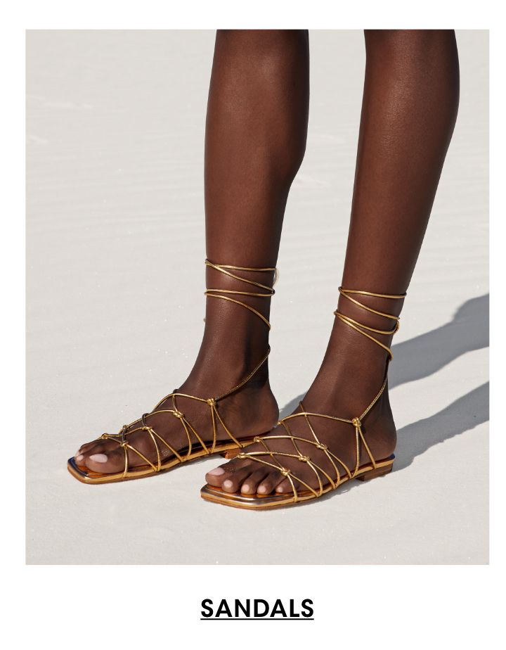 1 photo, a closeup of a female models legs and feet standing on sand, her feet in flat, strappy gold sandals that wrap up her ankles.