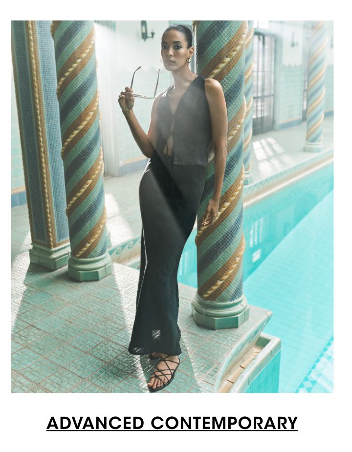 1 photo, of a female model leaning against a multicolor tiled column in an indoor pool, light coming through paneled doors. She holds sunglasses and wears a black vest and long black maxi skirt and black strappy flat sandals.