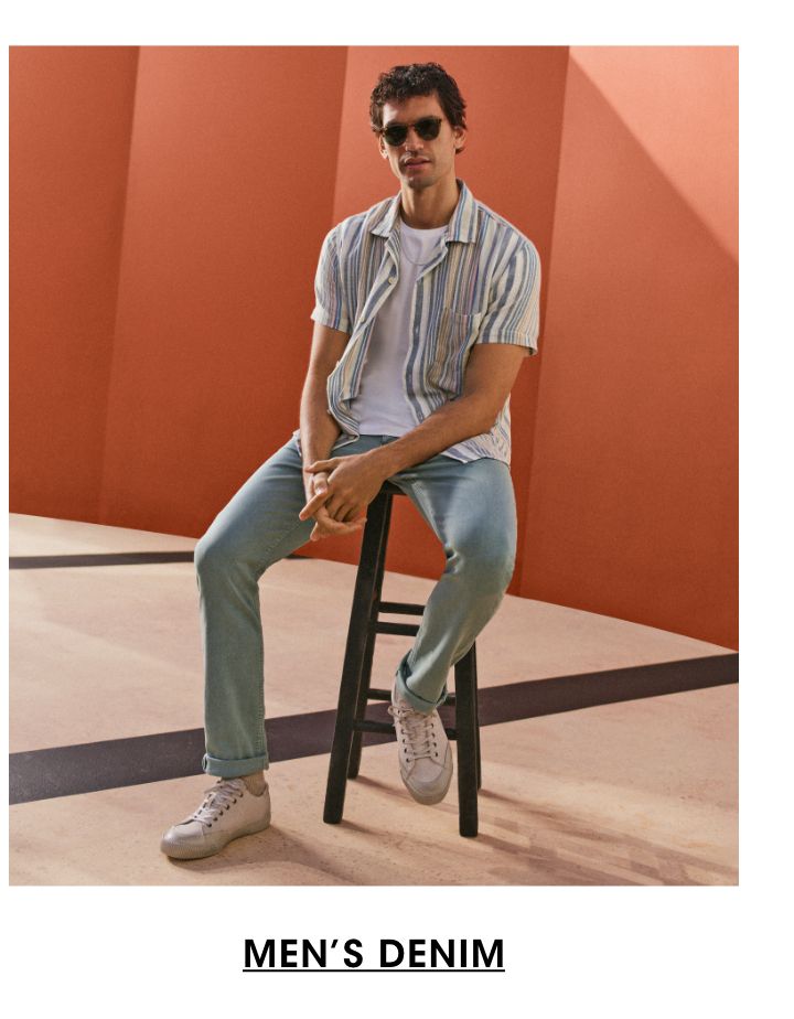 1 photo, of a male model sitting on a stool in front of an orange background. He wears sunglasses, a multicolored, striped, short sleeve unbuttoned buttoned down with a white tee shirt, light blue jeans, and white sneakers.