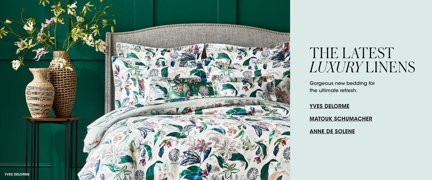 1 photo, of a bed with a brightly white and green plant printed duvet cover and pillows against a dark green wall, a round side table with 2 tall wicker vases with green flowers next to it.