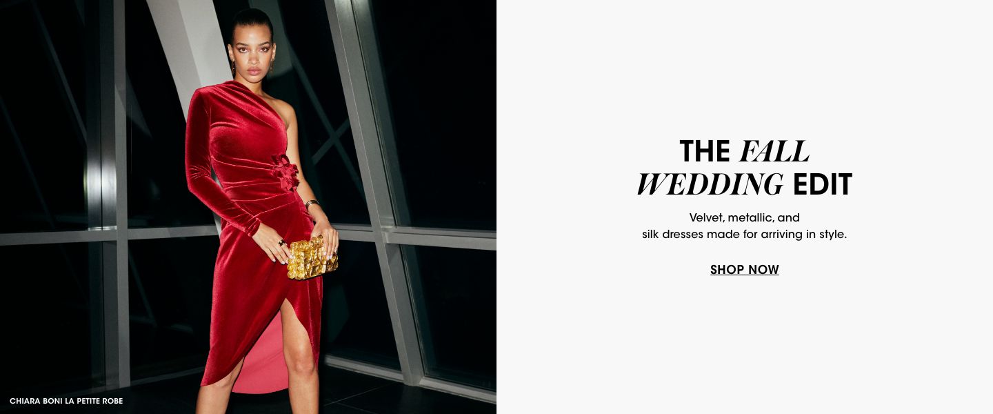 1 photo, of a female model standing at night in front of ceiling to floor glass windows, wearing a one sleeve velvet red dress and holding a gold clutch.