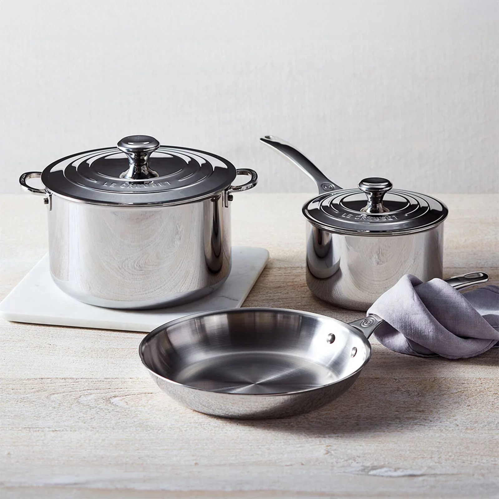 1 photo of 3 stainless steel Le Creuset cookware items, 1st of large pot with lid, 2nd of frying pan without lid, and 3rd saute pot with lid.