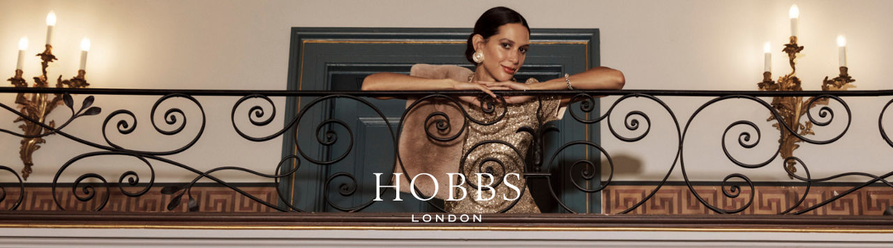 Hobbs London. A woman leaning on a railing dressed in a gold sparkling Hobbs London dress.