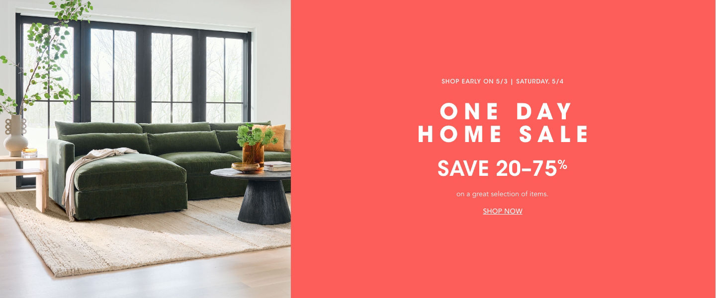 1 photo, of a green sectional couch in front of paneled floor to ceiling windows, a side table next to it with a vase with tall leafy branches, a round brown coffee table, all on a cream rug and light wood floor.