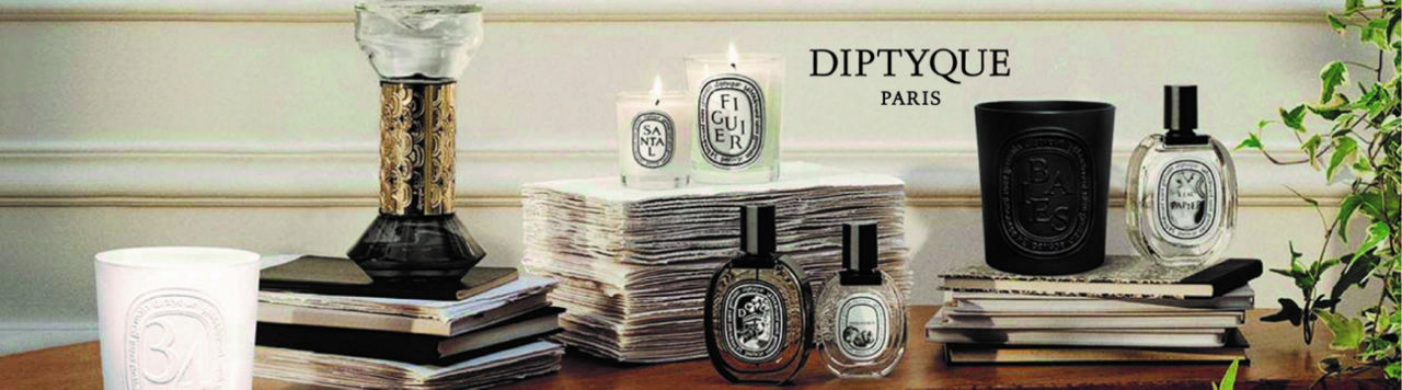 1 photo, of various Diptyque candles, some lit, and Diptyque perfume bottles arranged on a desk or on top of stacks of paper or books on the same table.