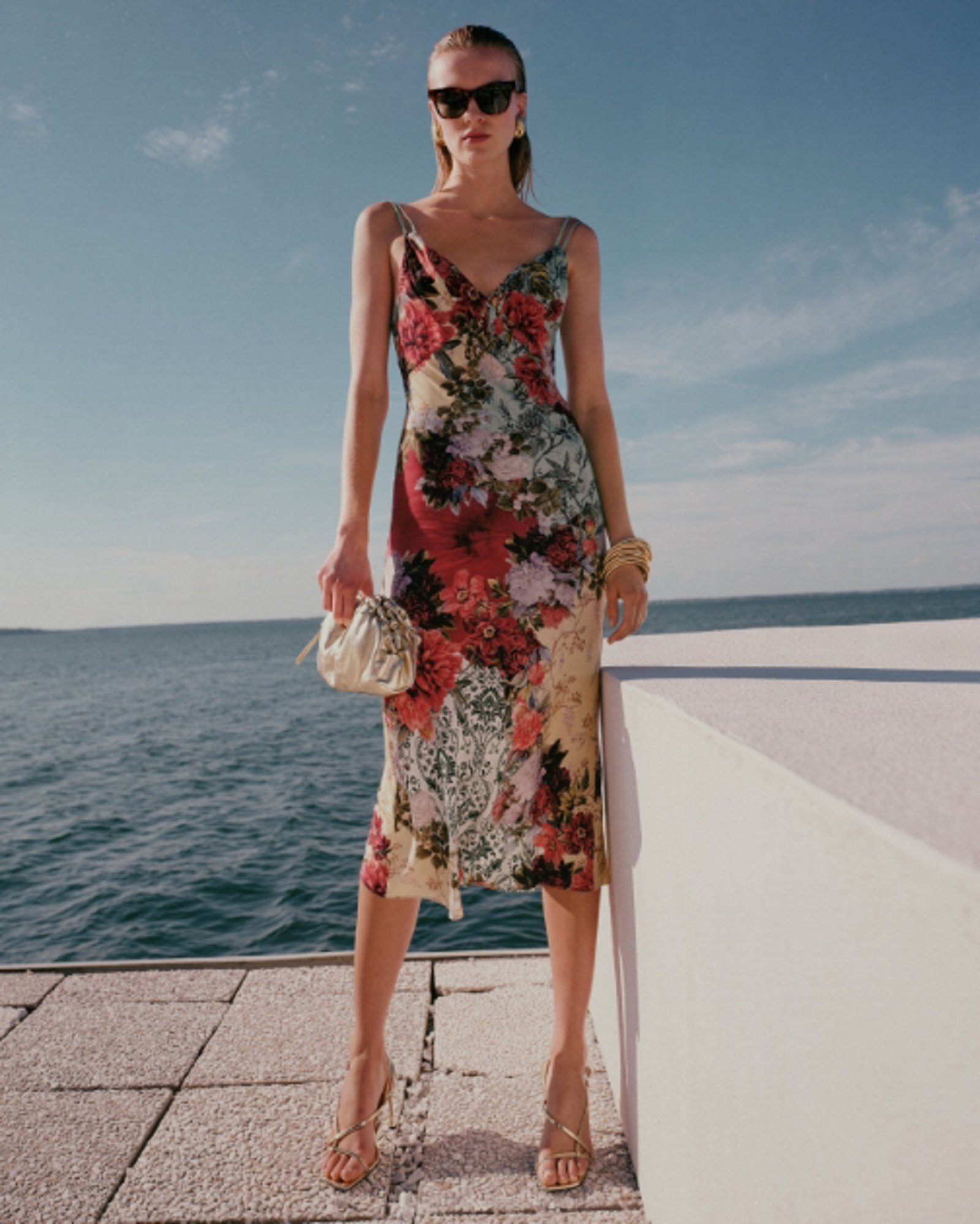 1 photo of female model in floral pattern cocktail dress, heels, sunglasses, and gold clutch.