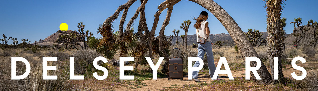 1 photo, of the back of a female model in a white top and jeans, a brown Delsey Paris roller luggage behind her, the model surrounded by desert Joshua trees.