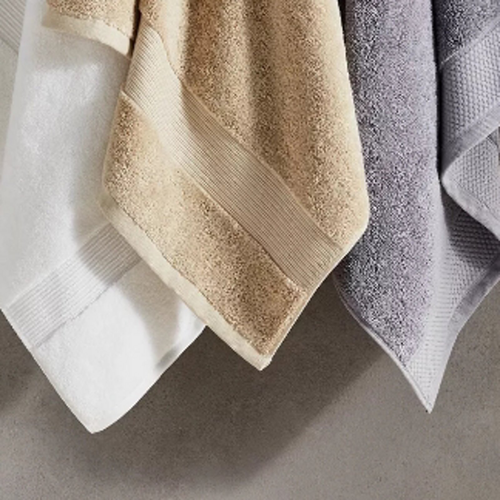 1 photo of 3 Hudson Park towels hanging against a wall, 1st in white, 2nd in beige, and 3rd in dark gray.