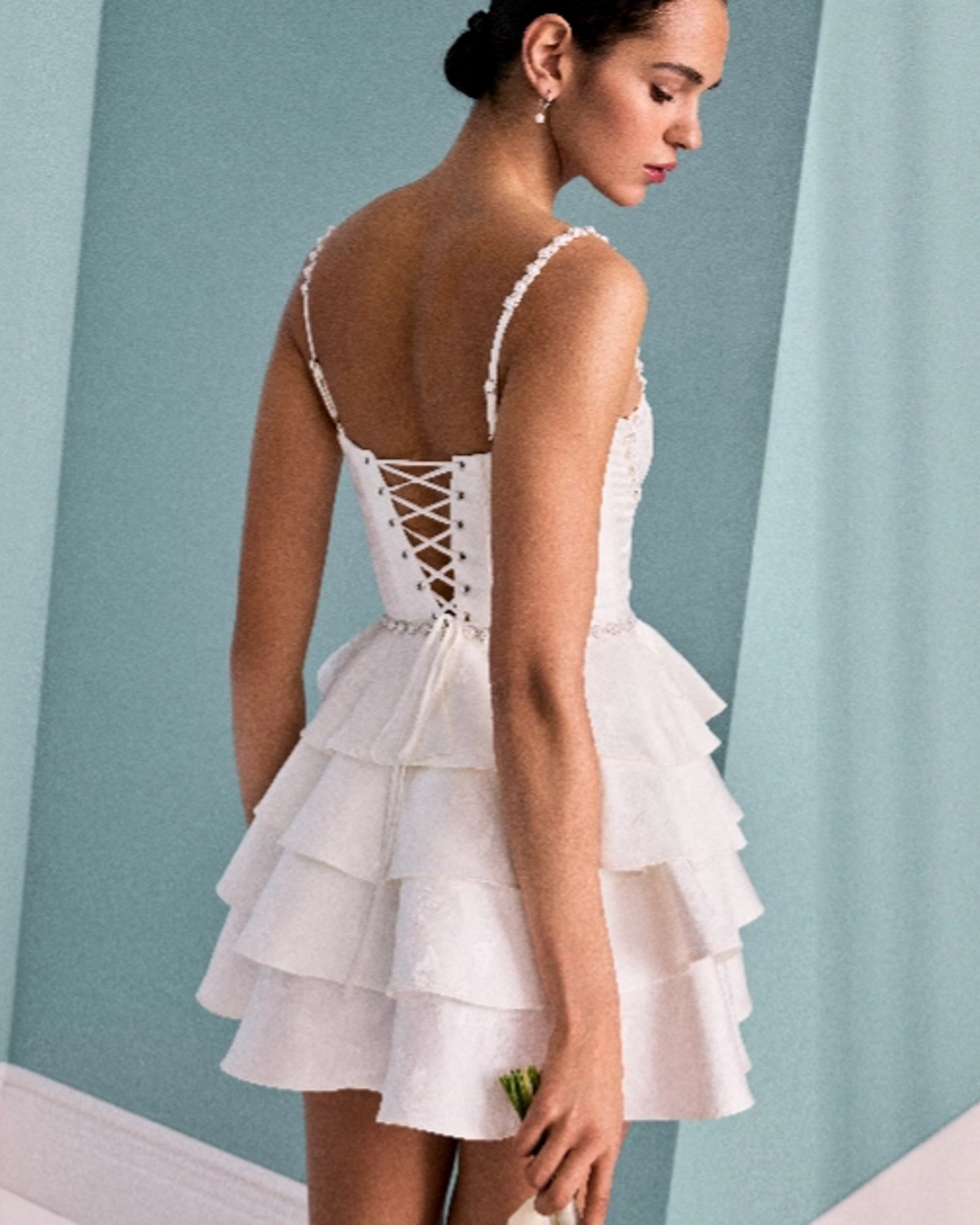 A woman from the back, wearing a white ruffled tiered mini dress with spaghetti straps and a corset tied back.