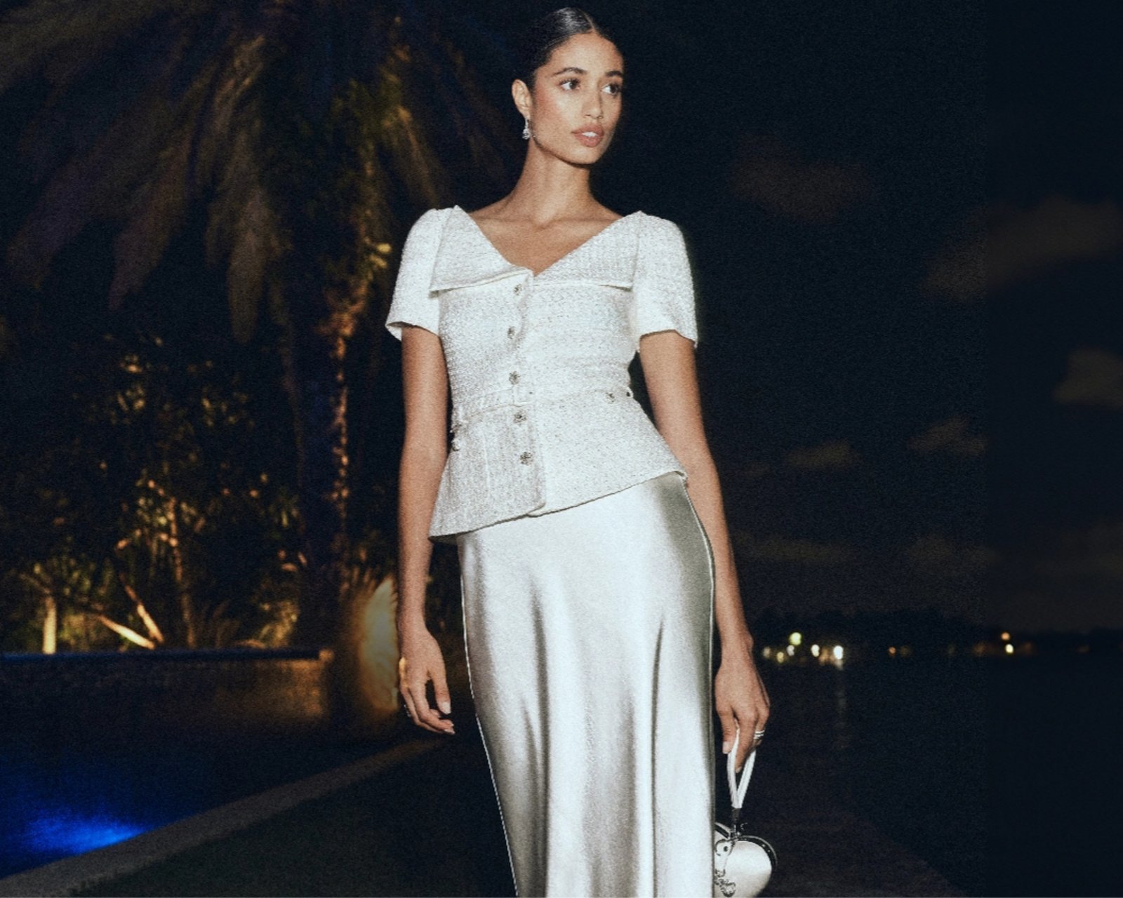 A woman at night, palm trees behind her, in a silver metallic button down short sleeve top and long satin skirt.