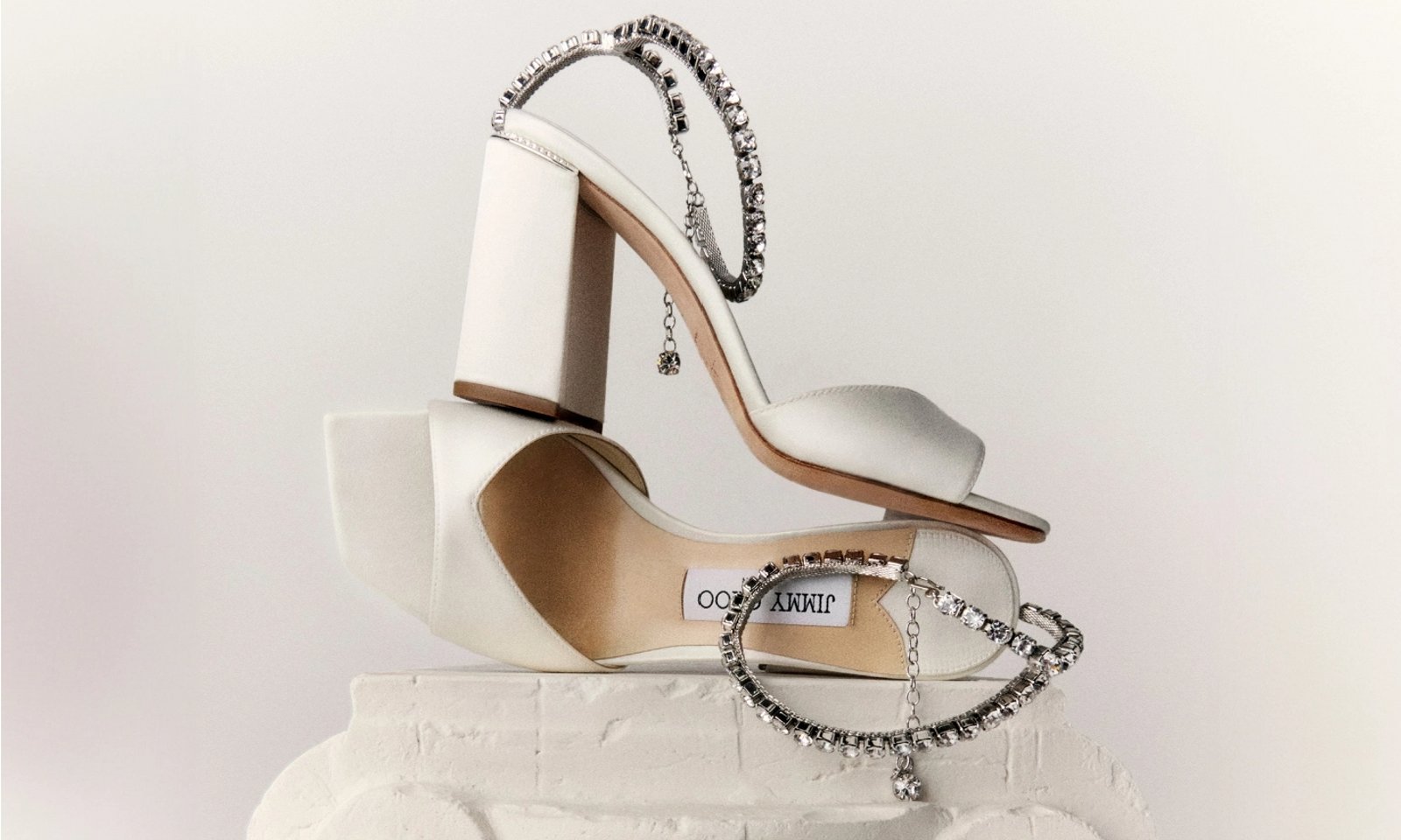 1 photo, of a pair of cream Jimmy Choo high heels with  embellished straps balanced atop each other on a short white cornice.