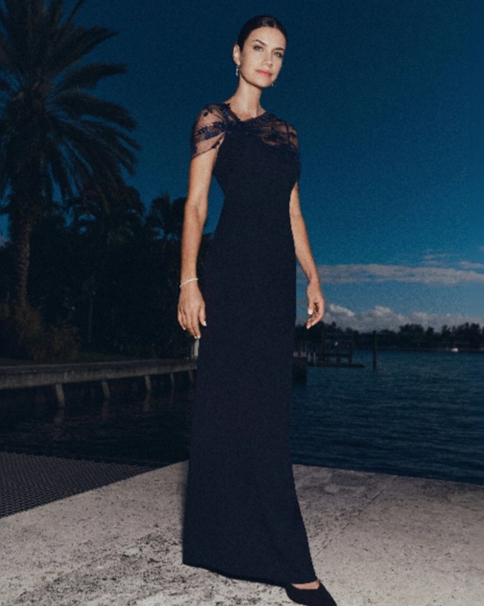 A woman standing outside with palm trees and water at dusk, wearing a long dark blue maxi dress with a mesh overlay over the shoulders.