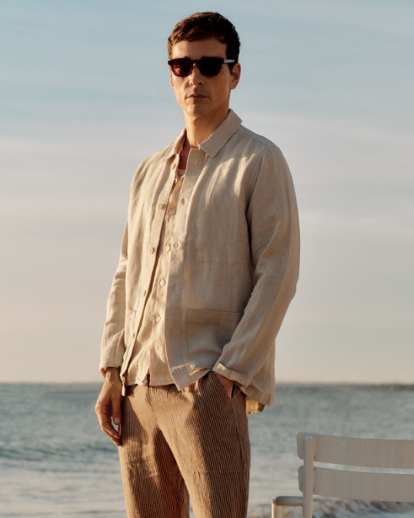 A man on a beach in a light brown linen button down and brown pants.