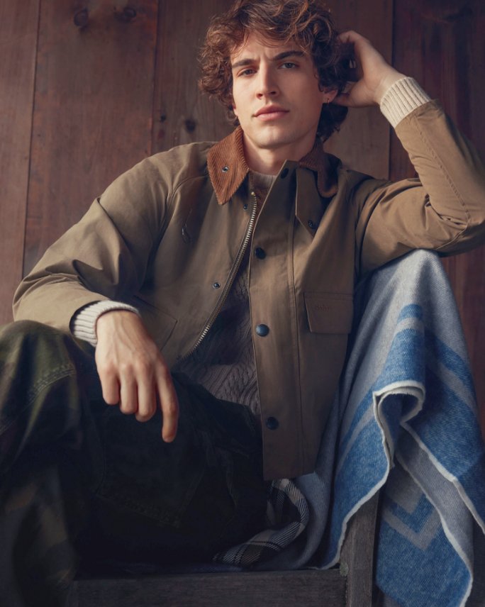 1 male model sitting in a chair in brown sweater, brown barn coat, and black pants.