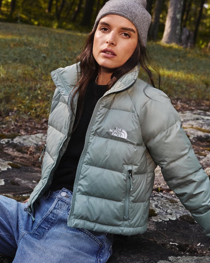 Female model in North Face cropped puffer coat in light green, black shirt, light blue jeans, and gray knitted ski hat.