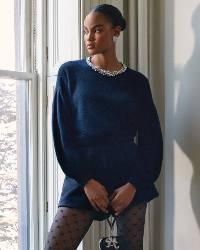 Female model in navy blue short knitted dress with pearl accents at the collar, navy blue sheer stockings and holding a black clutch.