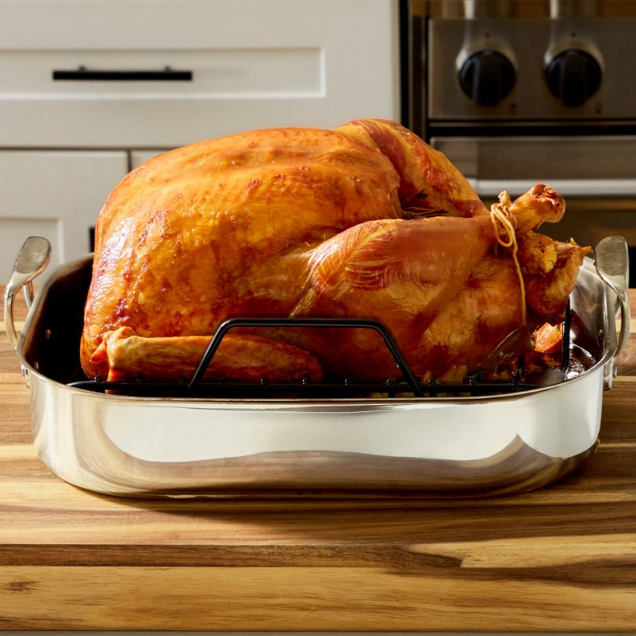 Stainless steel oven roaster with cooked turkey inside, set on top of a wooden counter in a kitchen.
