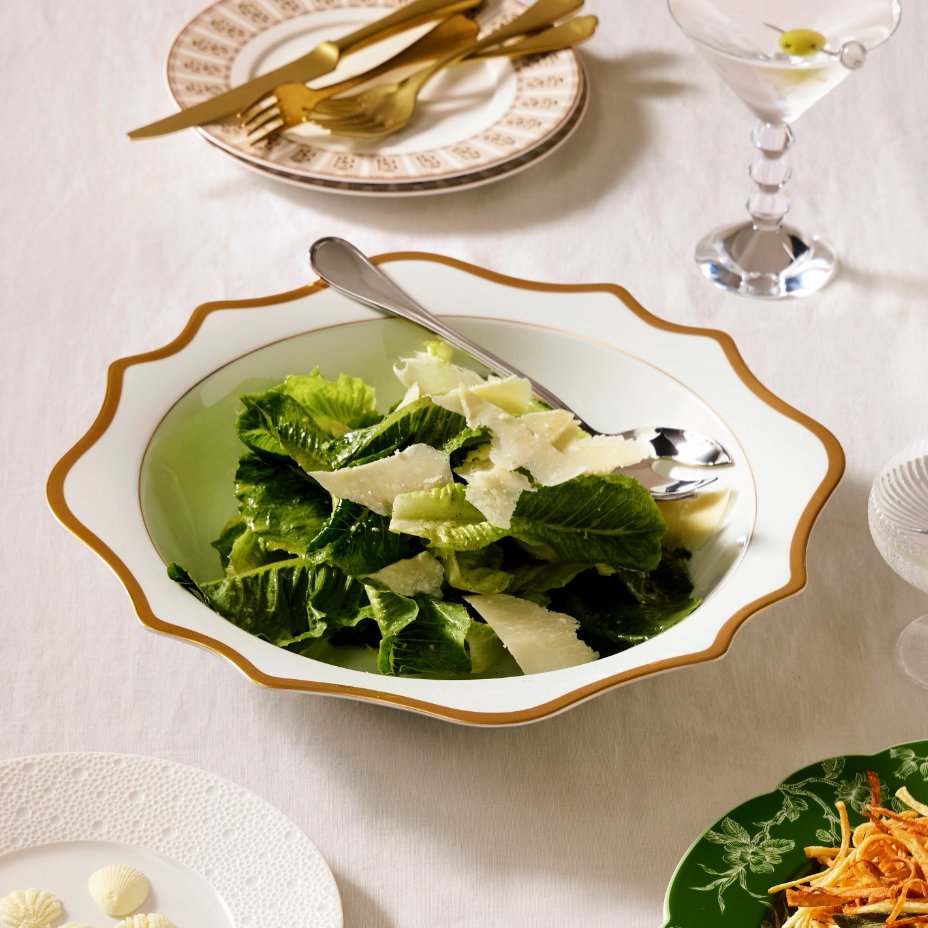 A white with gold trim salad bowl filled with a green salad on a white tabletop.