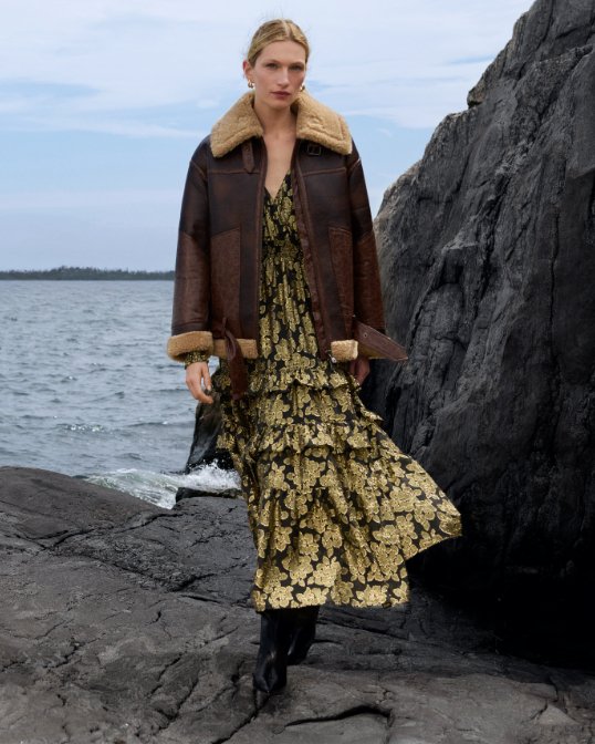 A woman on a rocky beach in a dark brown shearling lined barn jacket over a yellow and black printed dress.
