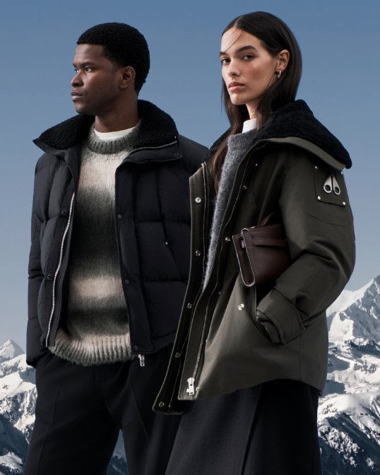 A man and woman, snowcapped mountains behind them, in black puffer coats.