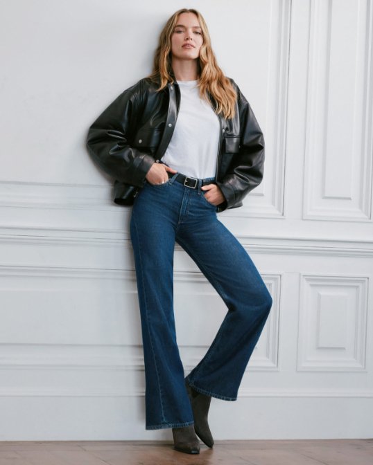 A woman leaning against a wall in a black jacket, white tee and dark blue jeans.