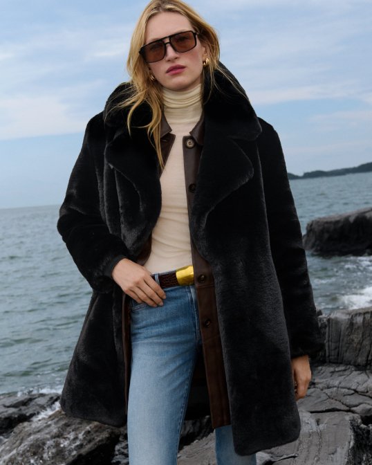 A woman on a rocky outcrop in a mid length brown faux fur jacket and blue jeans.