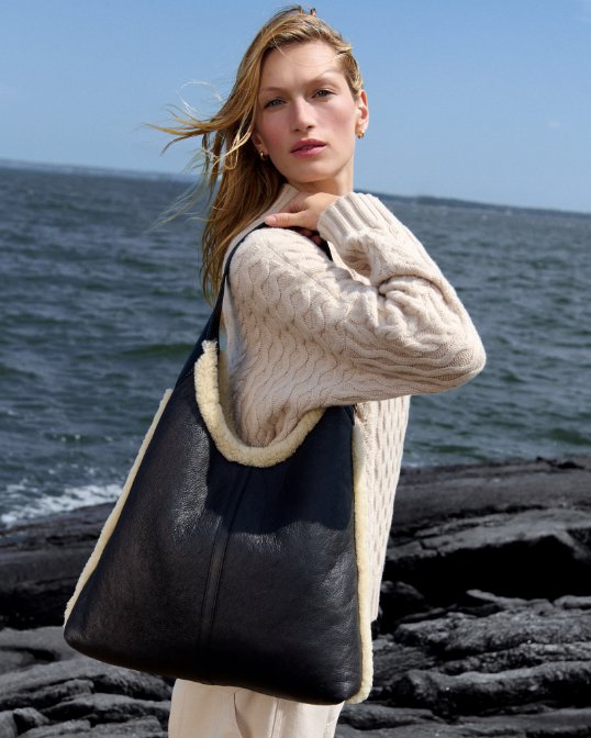 A woman in a cable knit sweater holding a large black tote with shearling lining.