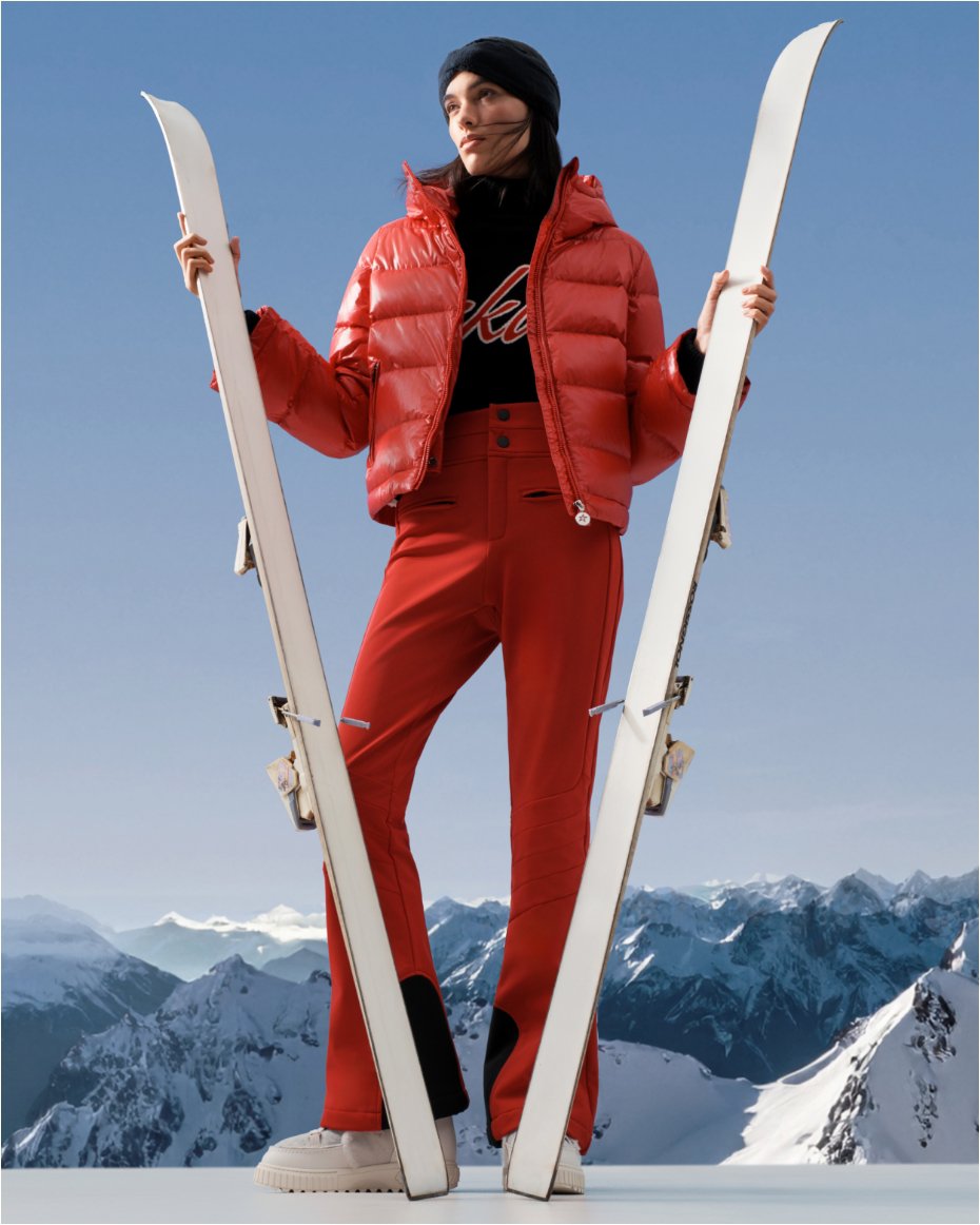 A woman on a snowy mountaintop in a red ski jacket and pants, holding upright a pair of skis.