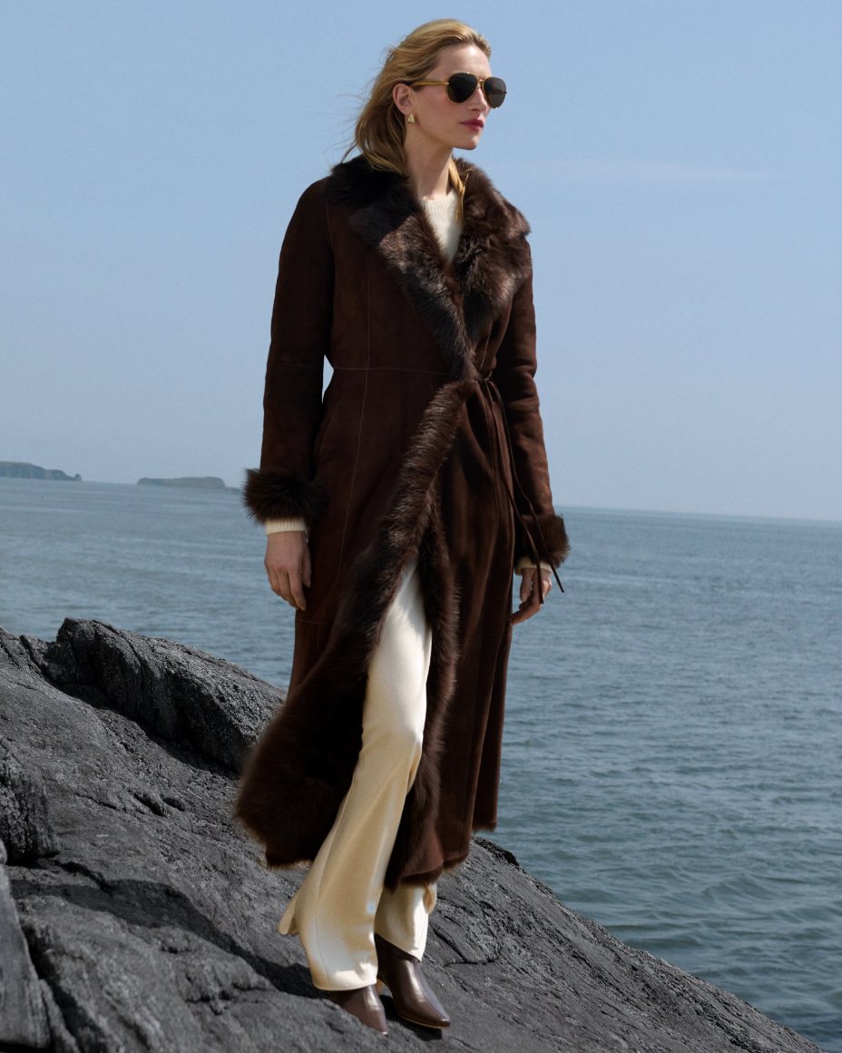 A woman standing on a rocky outcrop next to the ocean in a long brown faux fur coat. Shop the cold weather shop. 