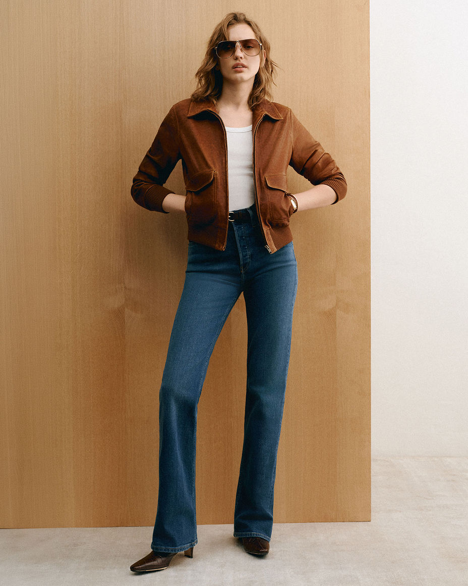 A woman in a brown suede jacket, white top & bootcut blue jeans. Shop bootcut jeans.