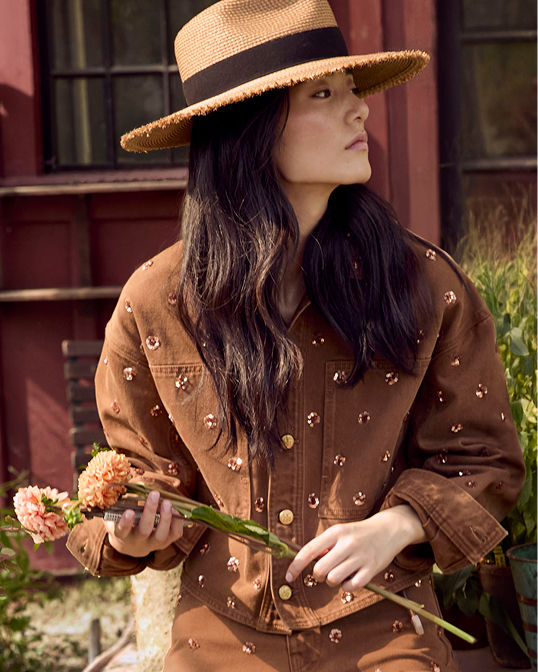 A woman in a brown embellished jacket and round brim hat. Shop coats and jackets.