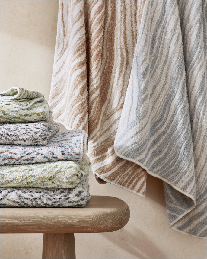 A folded pile of towels in a mix of patterns on a stool next to 2 more hanging towels.