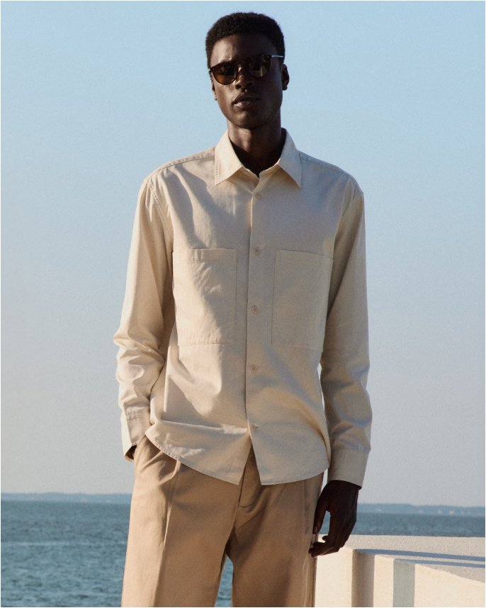 A man standing next to a lake in a beige button down and camel colored pants.