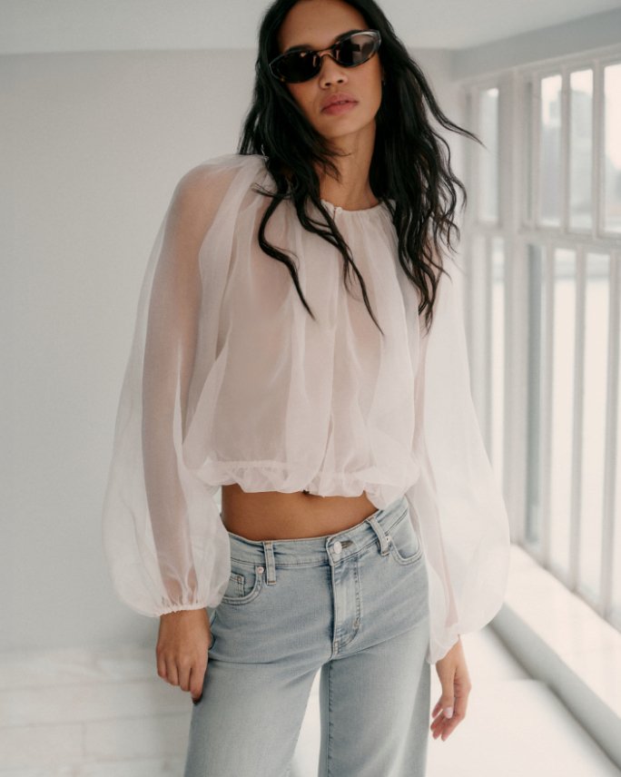 A woman in a white gauzy long sleeve top, light blue jeans, and sunglasses.