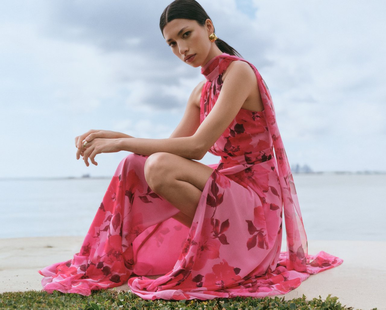 Woman in a pink halter dress with flower motif on a beach. Spring wedding season is starting. Shop wedding guest.