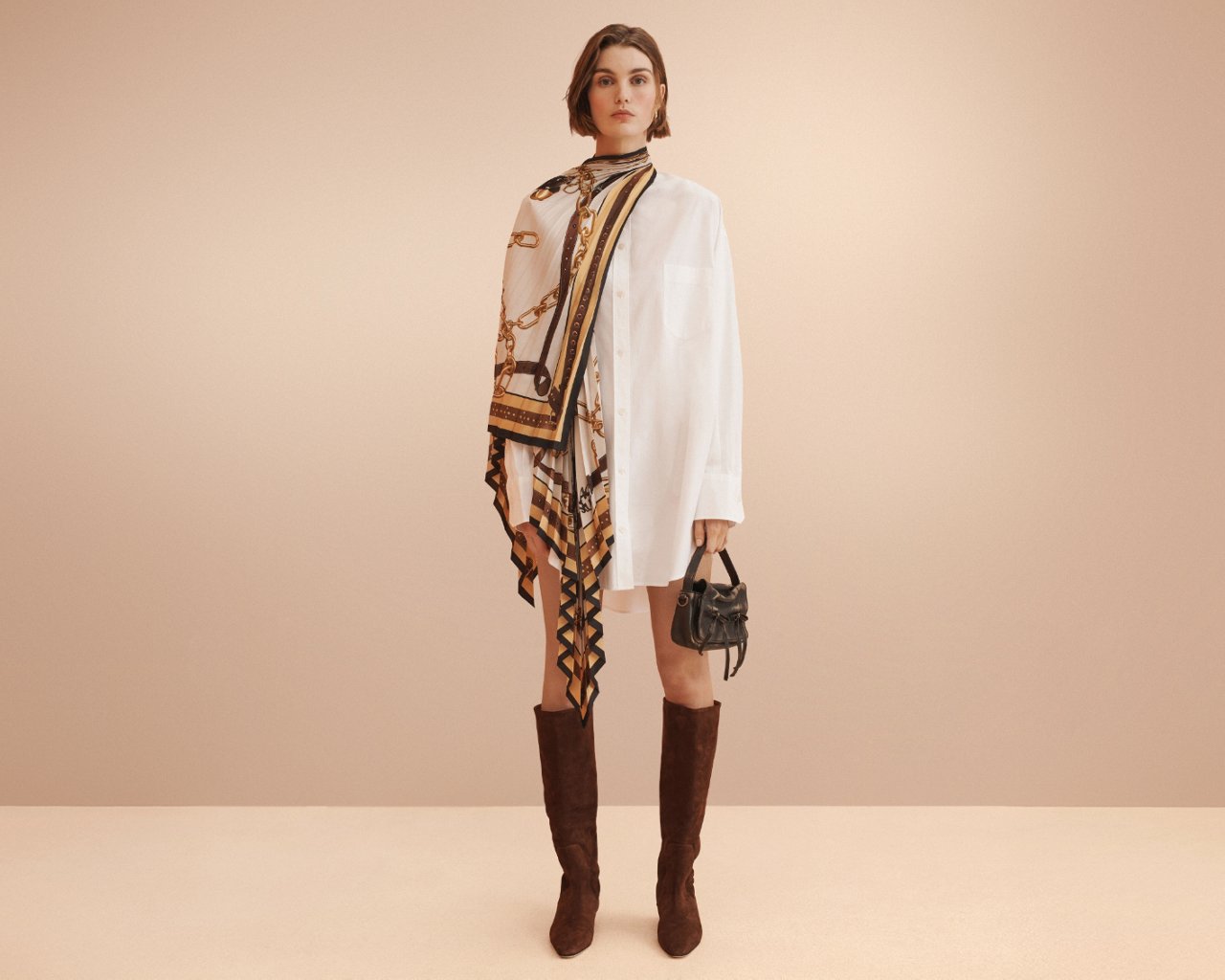 Woman in an Acne Studios white shirt dress with printed scarf across one shoulder and brown suede tall boots.