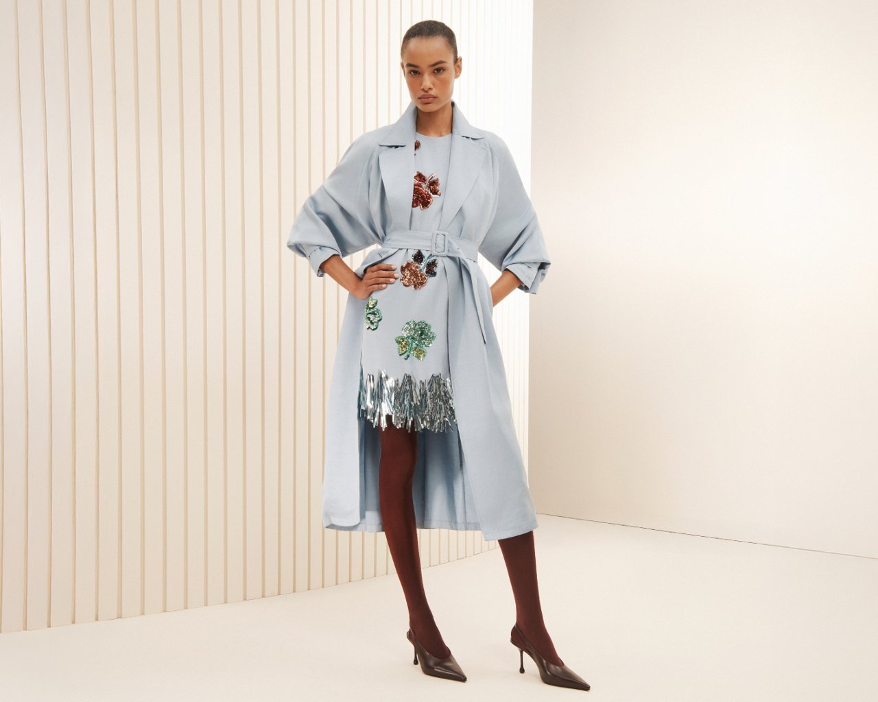 woman in a light blue Marni trench coat over a   sequined flower embroidered mini dress with silver hem.