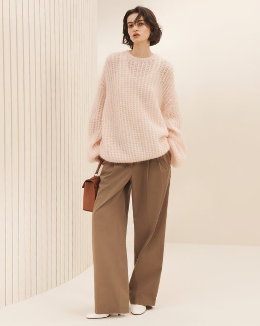 A female model in Toteme pale pink crewneck sweater, light brown trousers, and hold brown handbag.