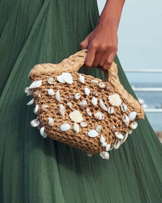 Woman in green skirt holding a brown crocheted handbag embellished with seashells.