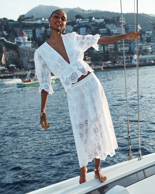 Woman balancing on the side of a boat in a white crocheted top & matching skirt.