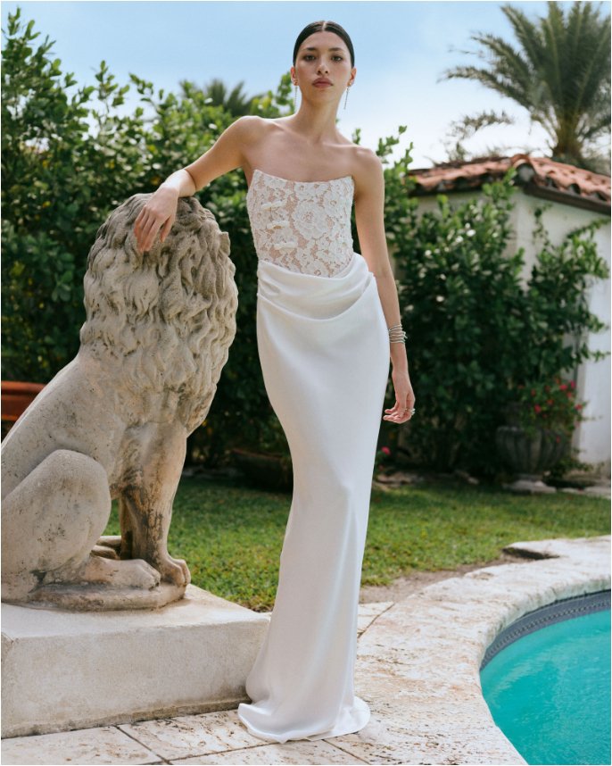 Woman leaning on stone lion in a formfitting strapless wedding dress with embroidered top. Shop featured shops, your bridal wardrobe.
