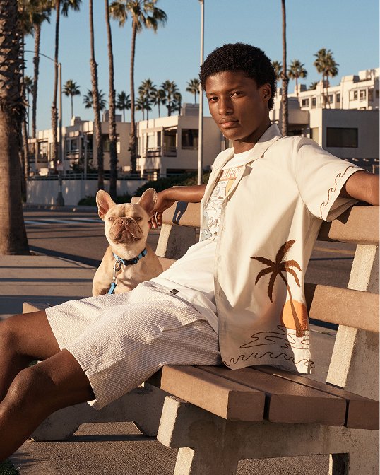 A man on a bench in a tropical print cream button down over a graphic tee and shorts.