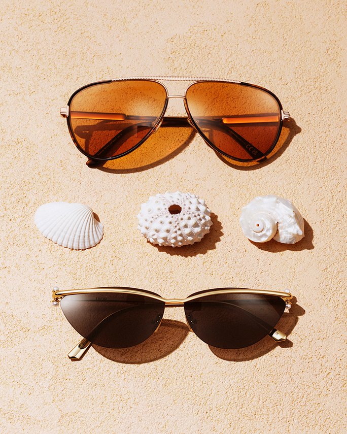 2 pairs of sunglasses on the sand, 3 shells lined up between them. Beach bound, sunglasses.