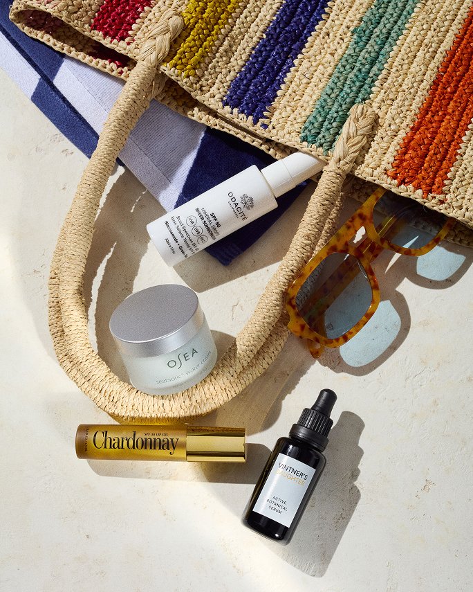 Beauty products & sunglasses spilling out of a multi color stripe woven tote. Beach bound, travel sized beauty.