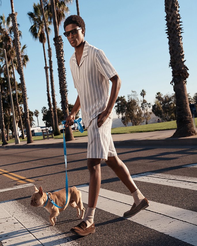 Man walking his dog across a street in a casual short sleeve striped shirt & matching shorts. Beach bound, vacation styles.