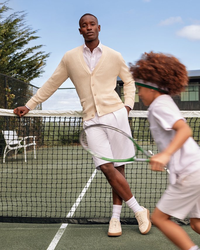 A young father on a tennis court in beige cardigan, white button down & shorts, his son running past with a racket. Fathers day gifts.