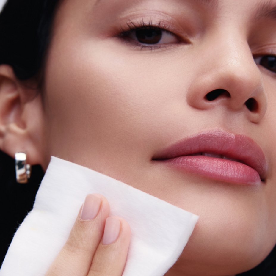 A womans face with minimal makeup, patting a makeup pad on her chin. Explore the guides, skincare.