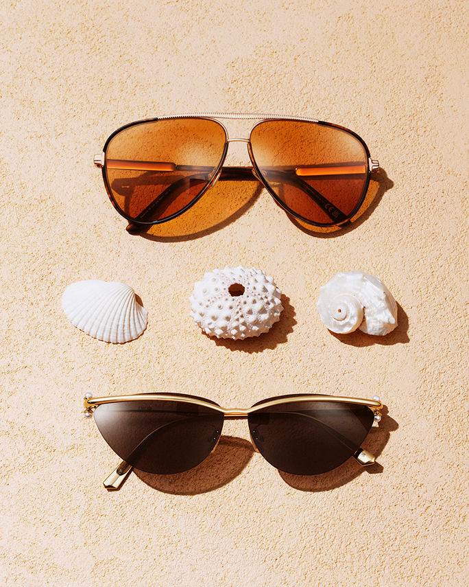 Two pairs of sunglasses on a sandy beach, sea shells between them. Featured shops, the vacation shop.